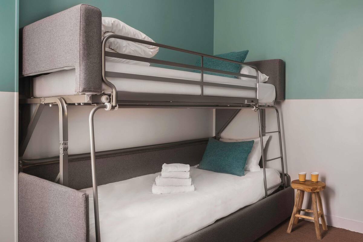 Una cama litera con estructura metálica, ropa de cama blanca y almohadas azul verdosas.