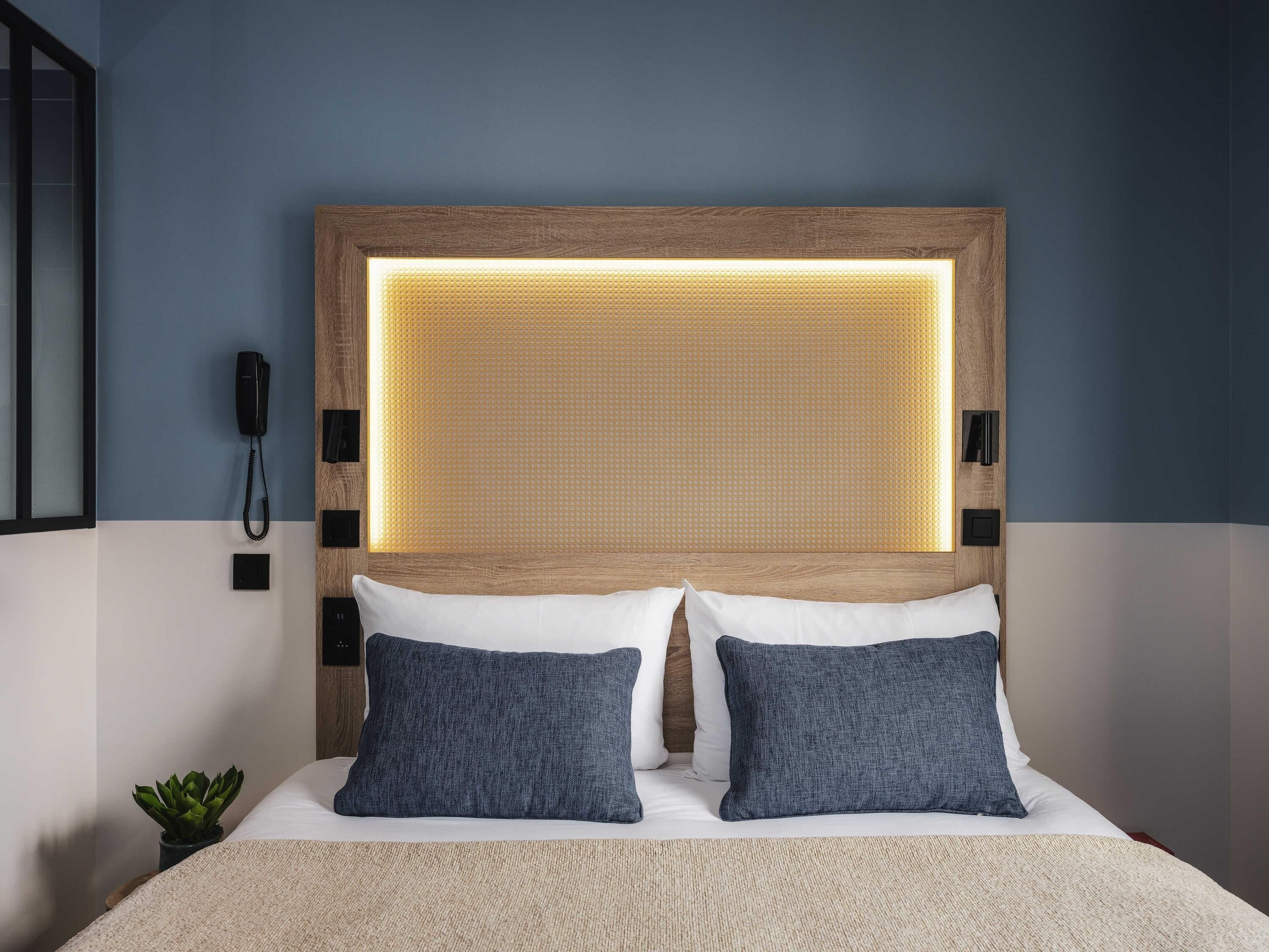 A hotel room with a bed, two pillows, a bedside phone, and a wall-mounted light fixture.