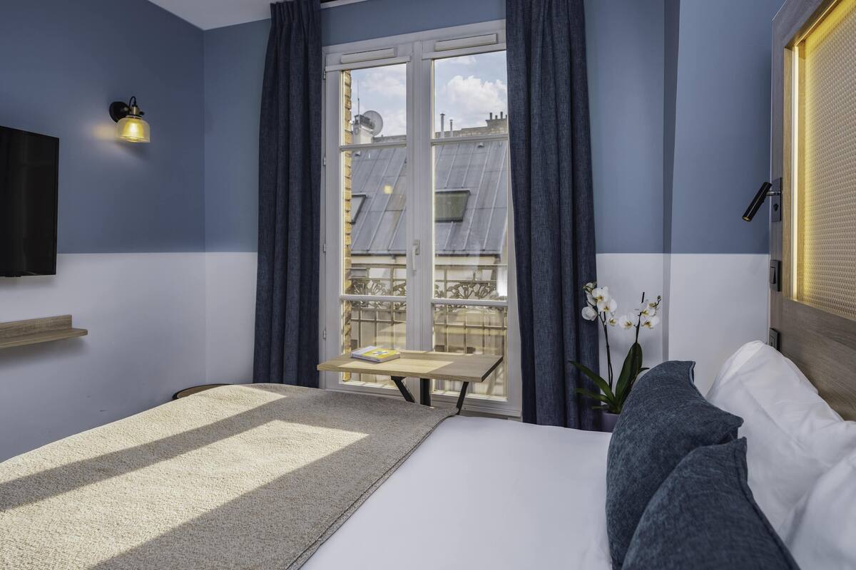 Une chambre à coucher comprenant un lit, une table de chevet, une lampe, une fenêtre avec des rideaux et une vue sur les bâtiments à l’extérieur.