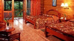 A wooden cabin with two beds, a dining area, and a balcony with a view of the forest.