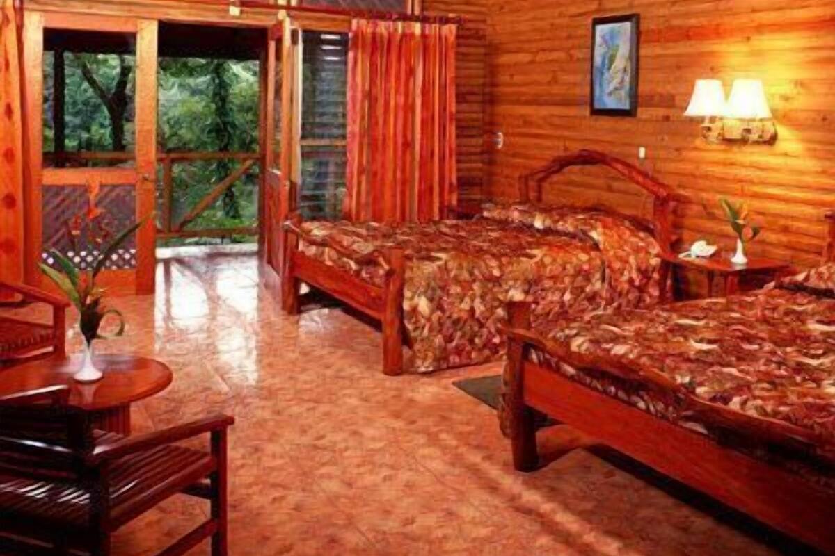 A wooden cabin with two beds, a dining area, and a balcony with a view of the forest.