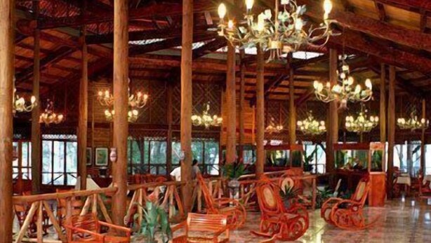 A spacious hall with high wooden beams and chandeliers, furnished with wicker chairs and tables, and large windows allowing natural light.