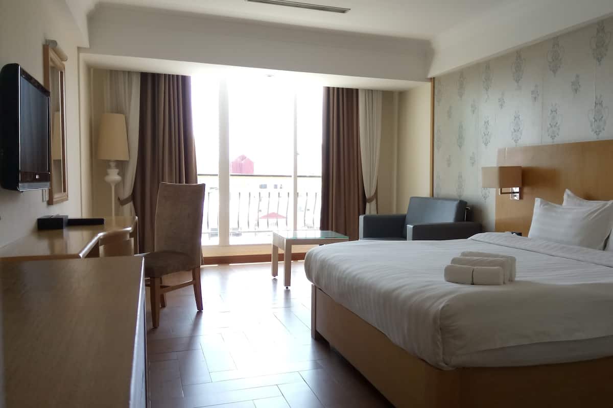 A hotel room with a large bed, a desk, a chair, a TV, and a balcony with curtains.