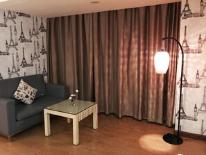 A room with a blue sofa, a glass coffee table, a floor lamp, and curtains with a Paris-themed wallpaper.