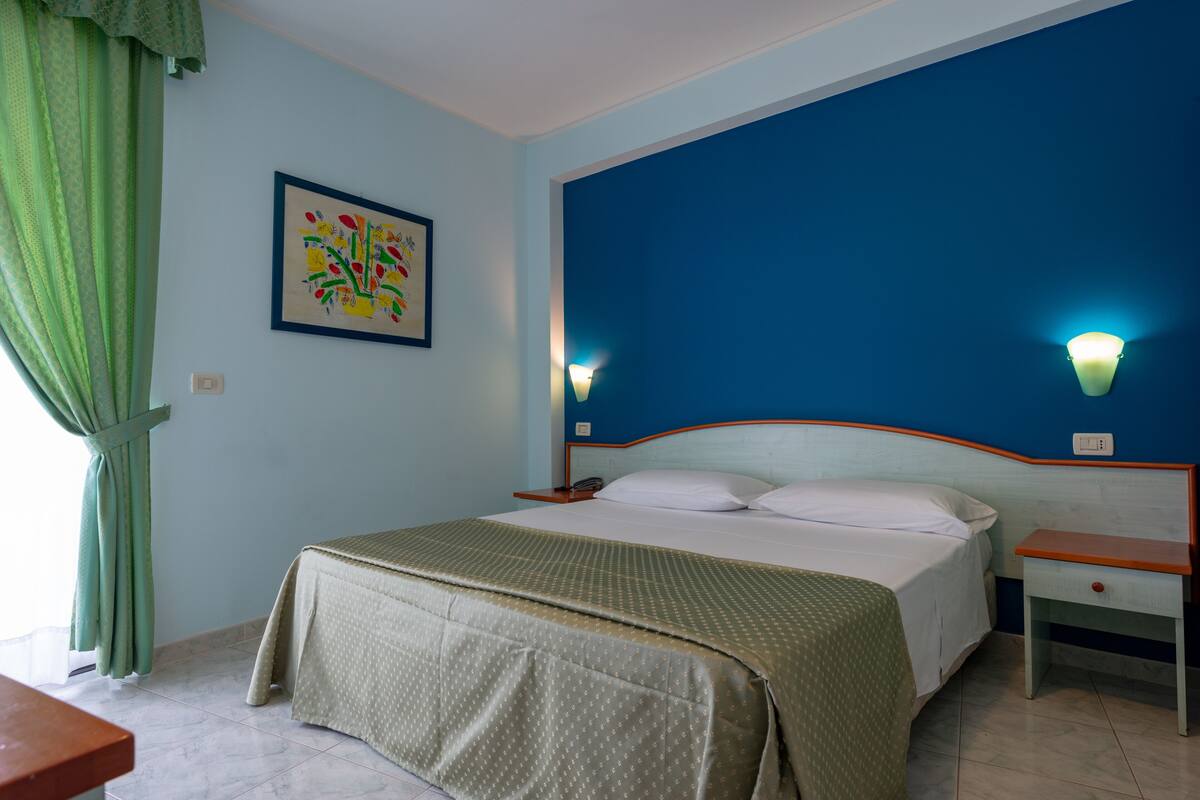 A hotel room with a bed, a nightstand, a bedside table, a wall-mounted light, a window with curtains, and a framed picture on the wall.