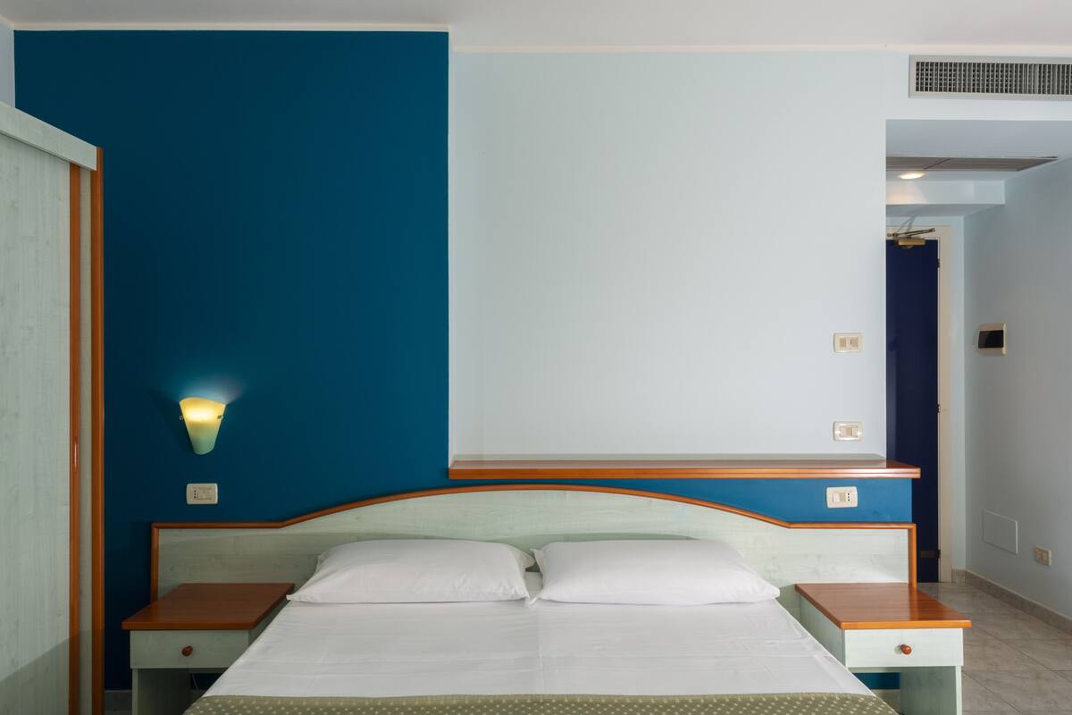 A hotel room with a bed, wooden headboard, nightstands, and a wall-mounted light.