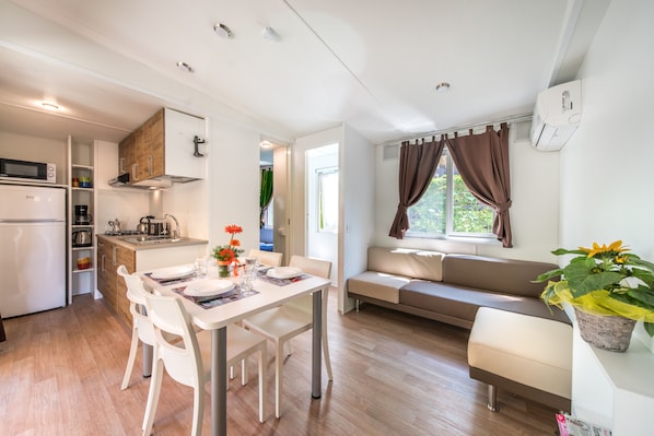 Una cocina moderna con armarios blancos, microondas y refrigerador. Hay una zona de comedor con una mesa y sillas blancas, y un sofá con vista a la ventana.