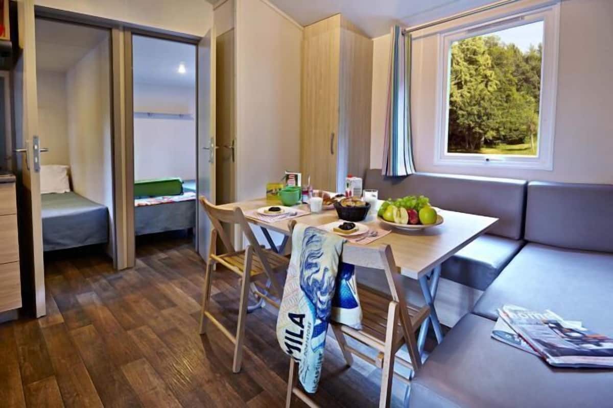 A compact room with a dining table, chairs, a bench, a bed, and a window with a view of trees.