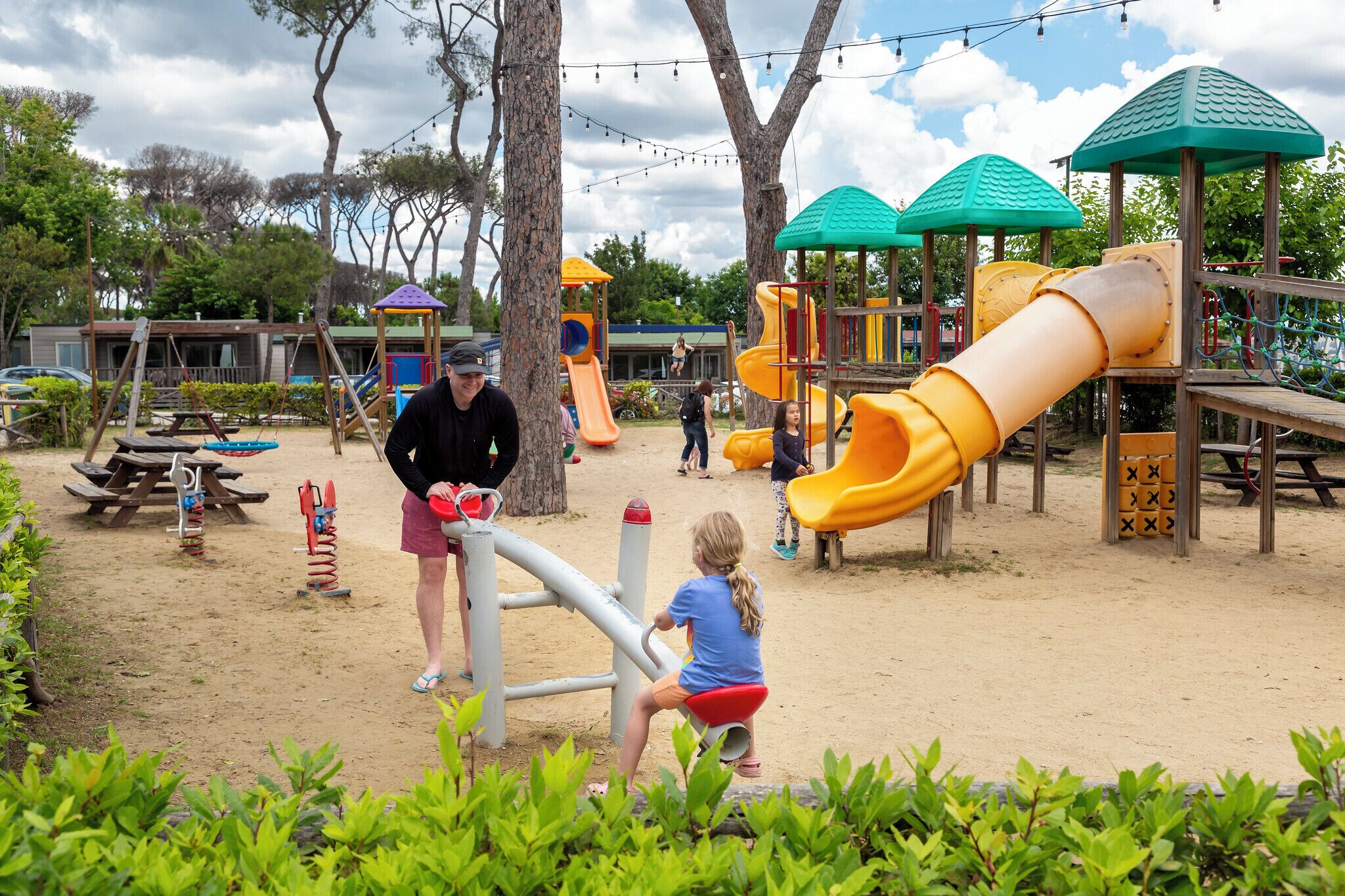 Ein Spielplatz mit verschiedenen Spielgeräten, darunter Rutschen und Kletterbereichen, umgeben von Bäumen und Picknicktischen.