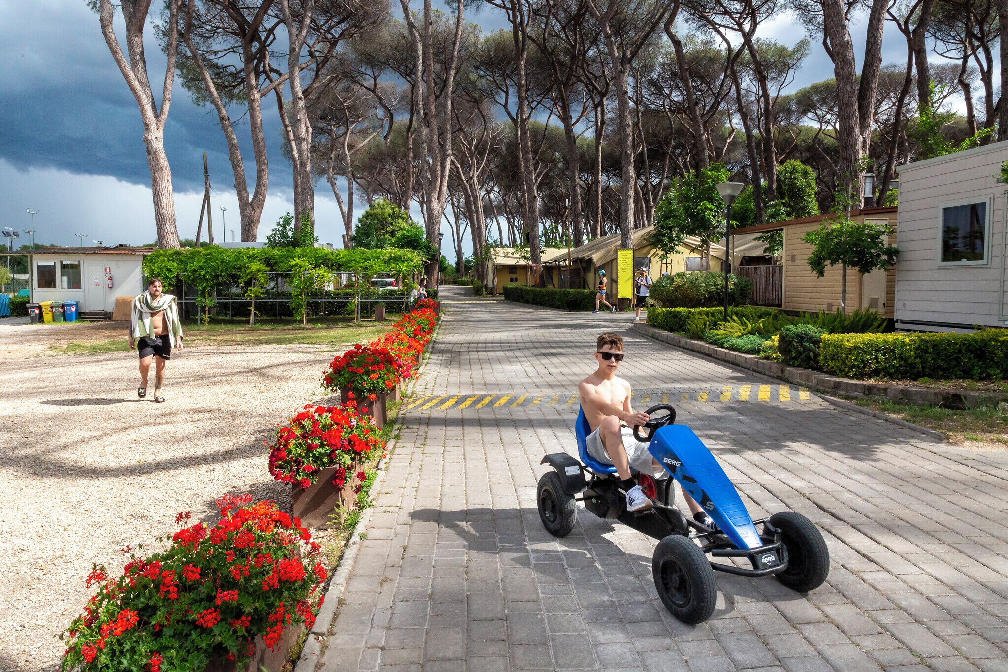 A paved pathway lined with red flowers, a person on a blue go-kart, and mobile homes in the background.
