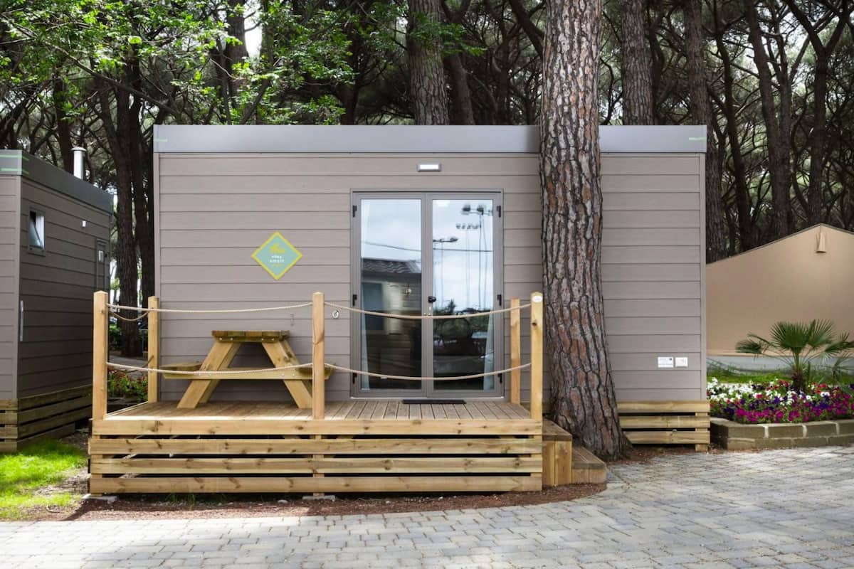 A small wooden structure with a bench and a glass door, surrounded by trees and a paved area.