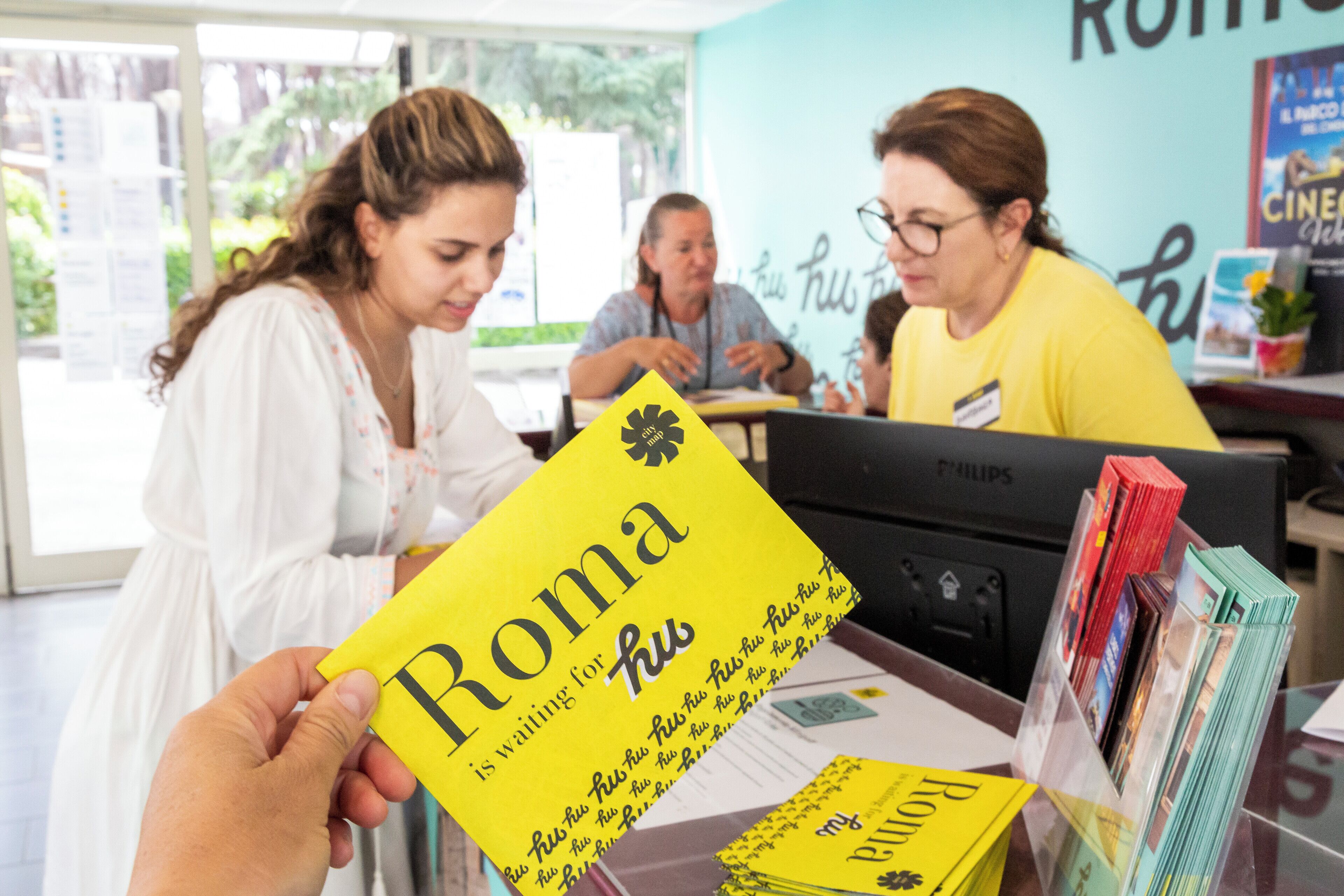 A person holding a yellow book titled "Roma" in a room with a table, a computer, and a magazine rack.