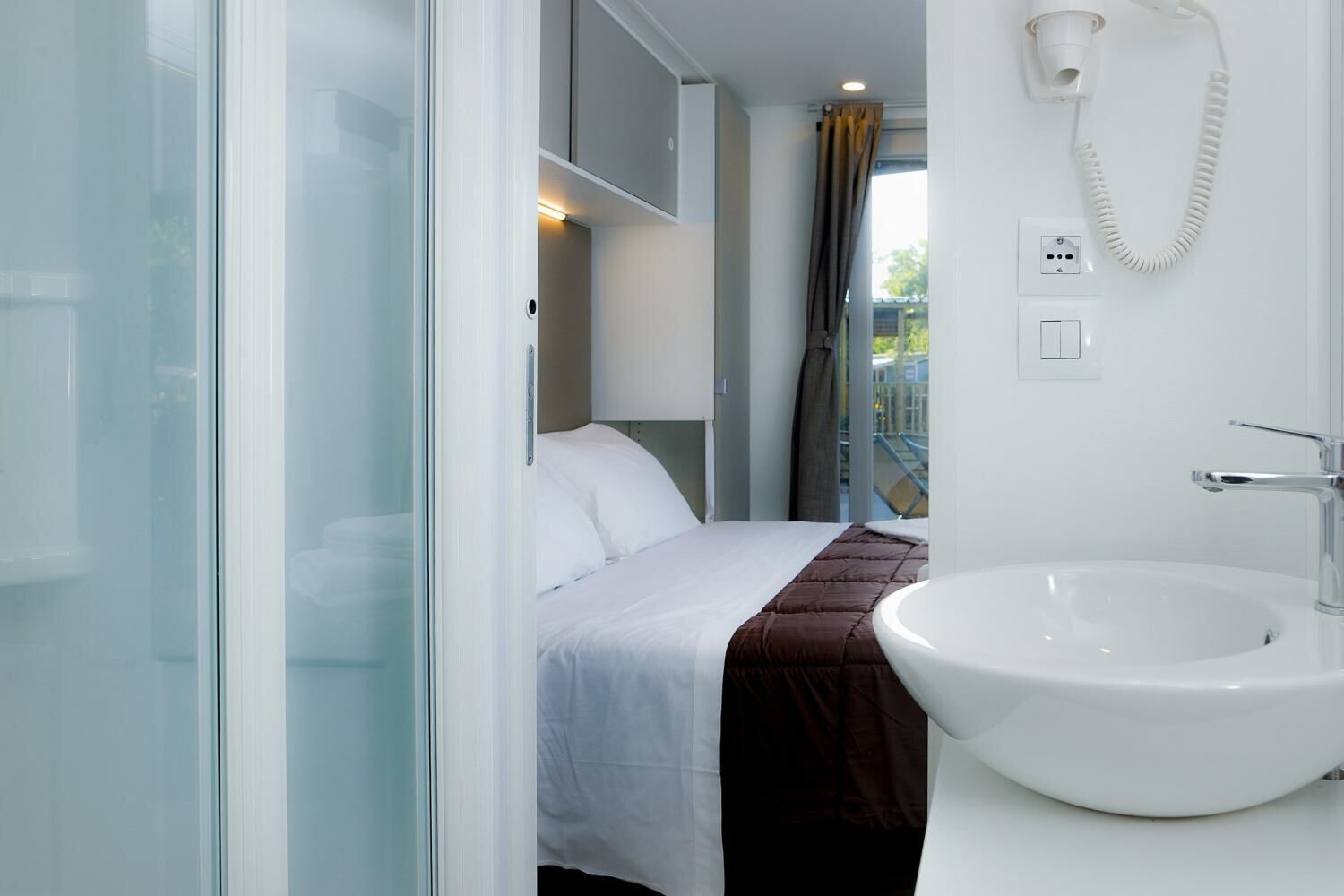 A modern hotel room with a bed, a glass shower enclosure, a white sink, and a telephone.