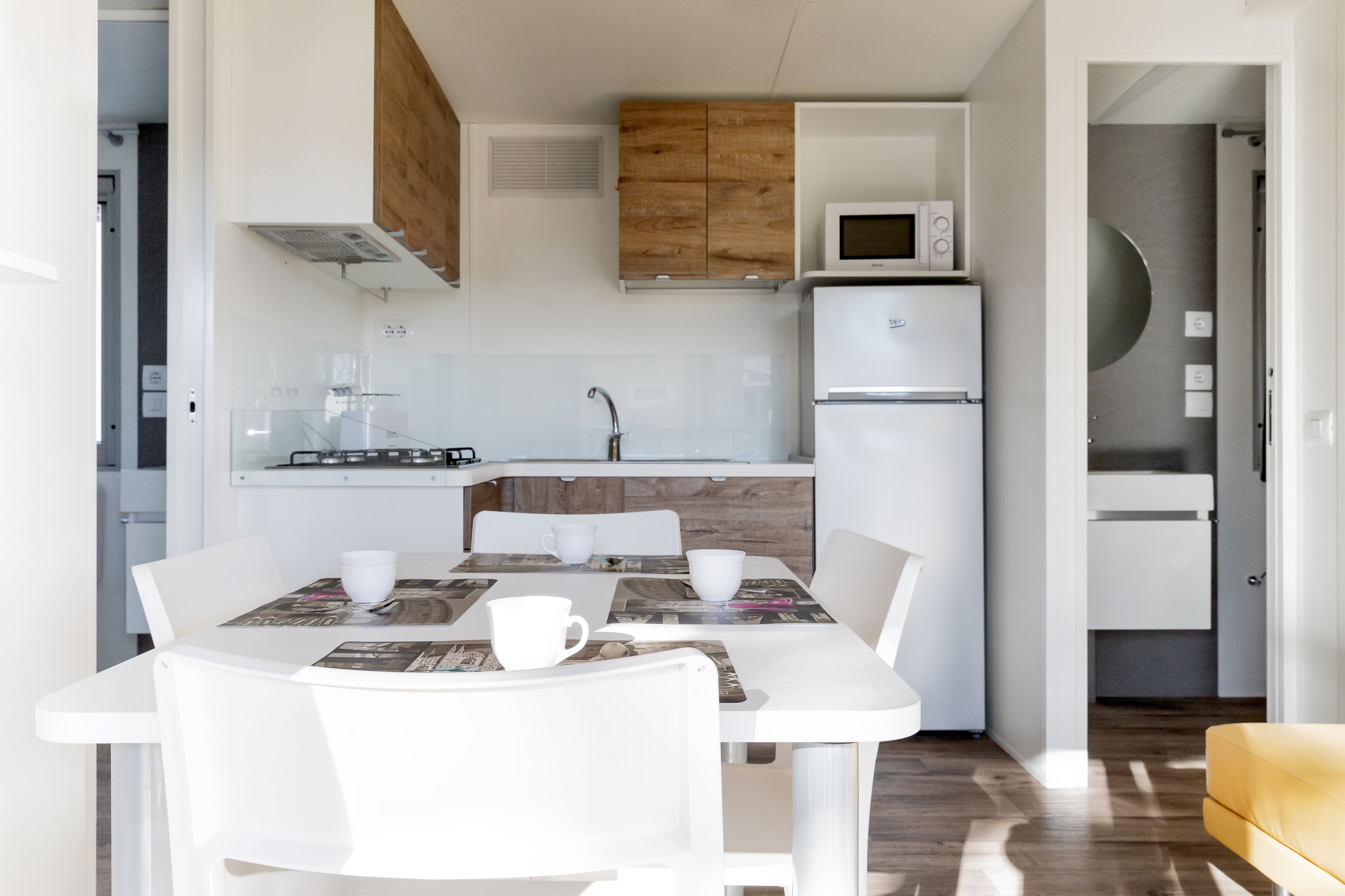 Een compacte keuken met witte apparaten, een eettafel met stoelen en een gele bank.