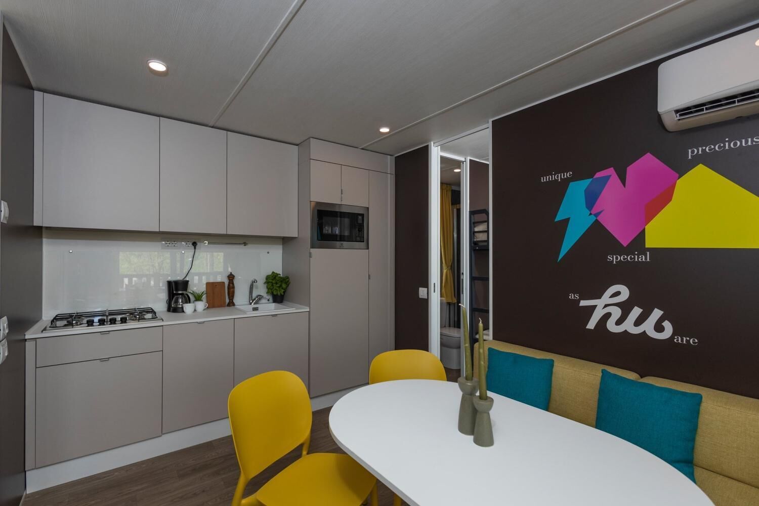 A modern kitchen with grey cabinets, a white countertop, and a stove. There is a dining area with a white table and yellow chairs. A wall with colorful geometric shapes and text is visible.