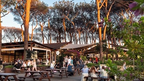 An outdoor dining area with wooden tables and benches, surrounded by trees and a building with large windows. - Fabulous Village (Rome)