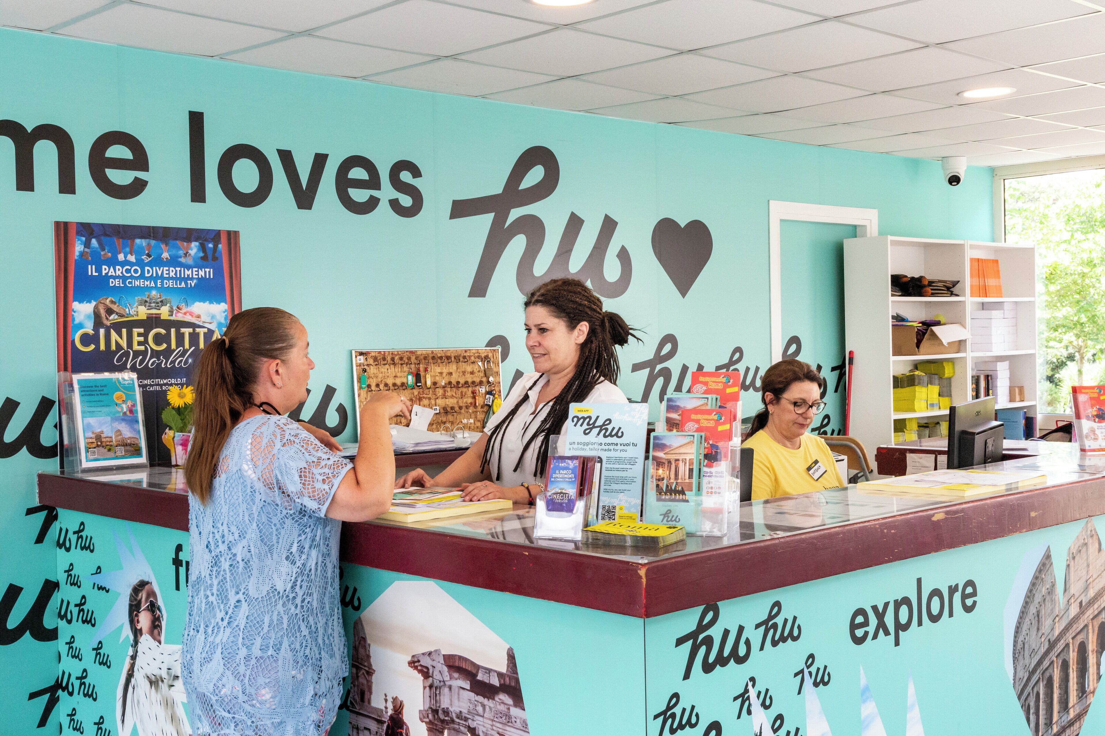 A reception desk with a teal wall, a poster of 'Cinecitt' and 'Il Parco Divertimenti', and a sign that reads 'hu hu explore'.