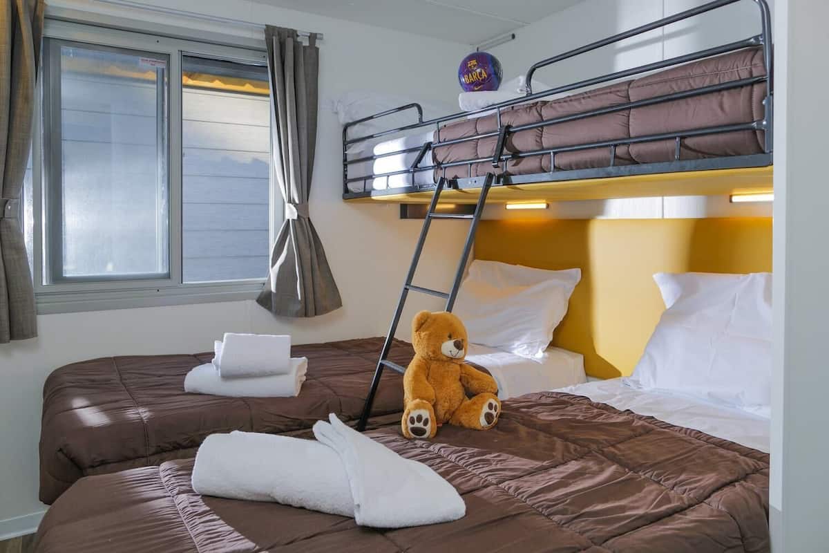 A hotel room with a bunk bed, a teddy bear on the lower bed, and a window with curtains.