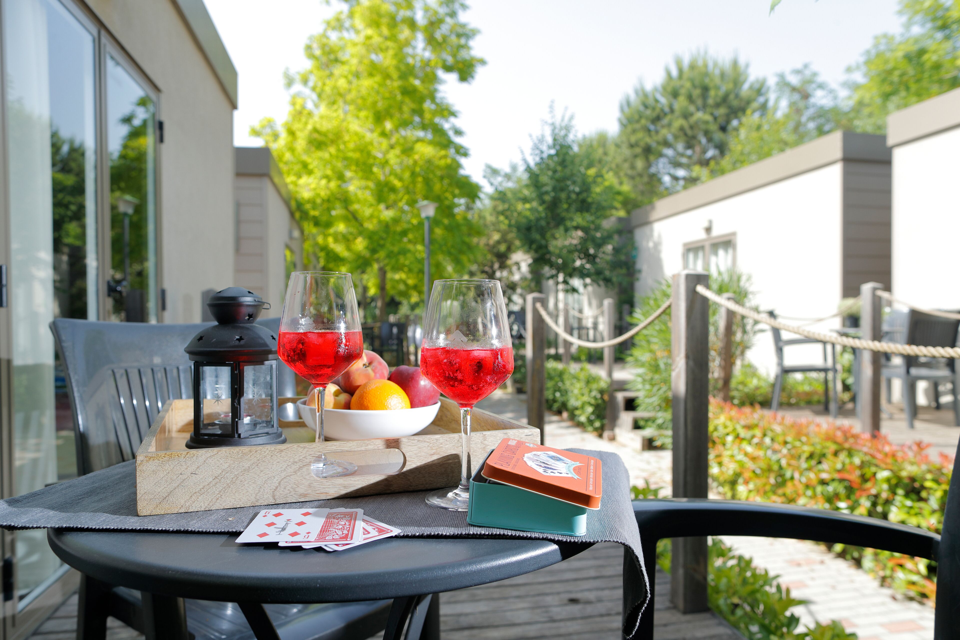 Una mesa con una bandeja de fruta, dos vasos de bebida roja, un farol y una caja pequeña en una terraza.