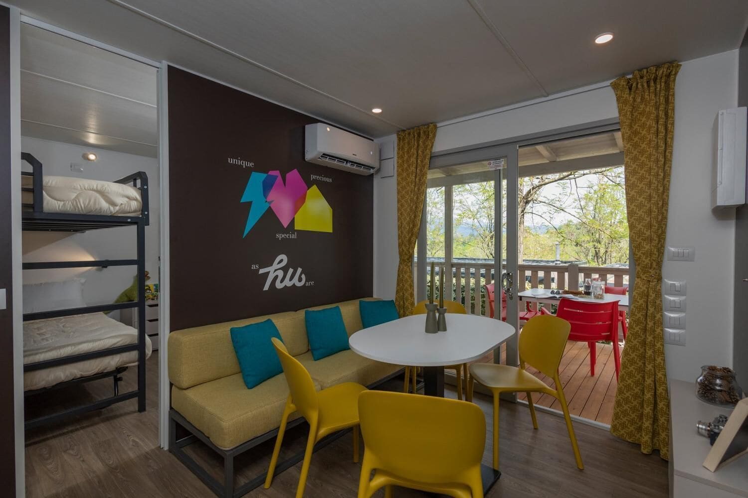 A room with bunk beds, a sofa, a round table, and yellow chairs. There is a window with curtains and a view of the outdoors.