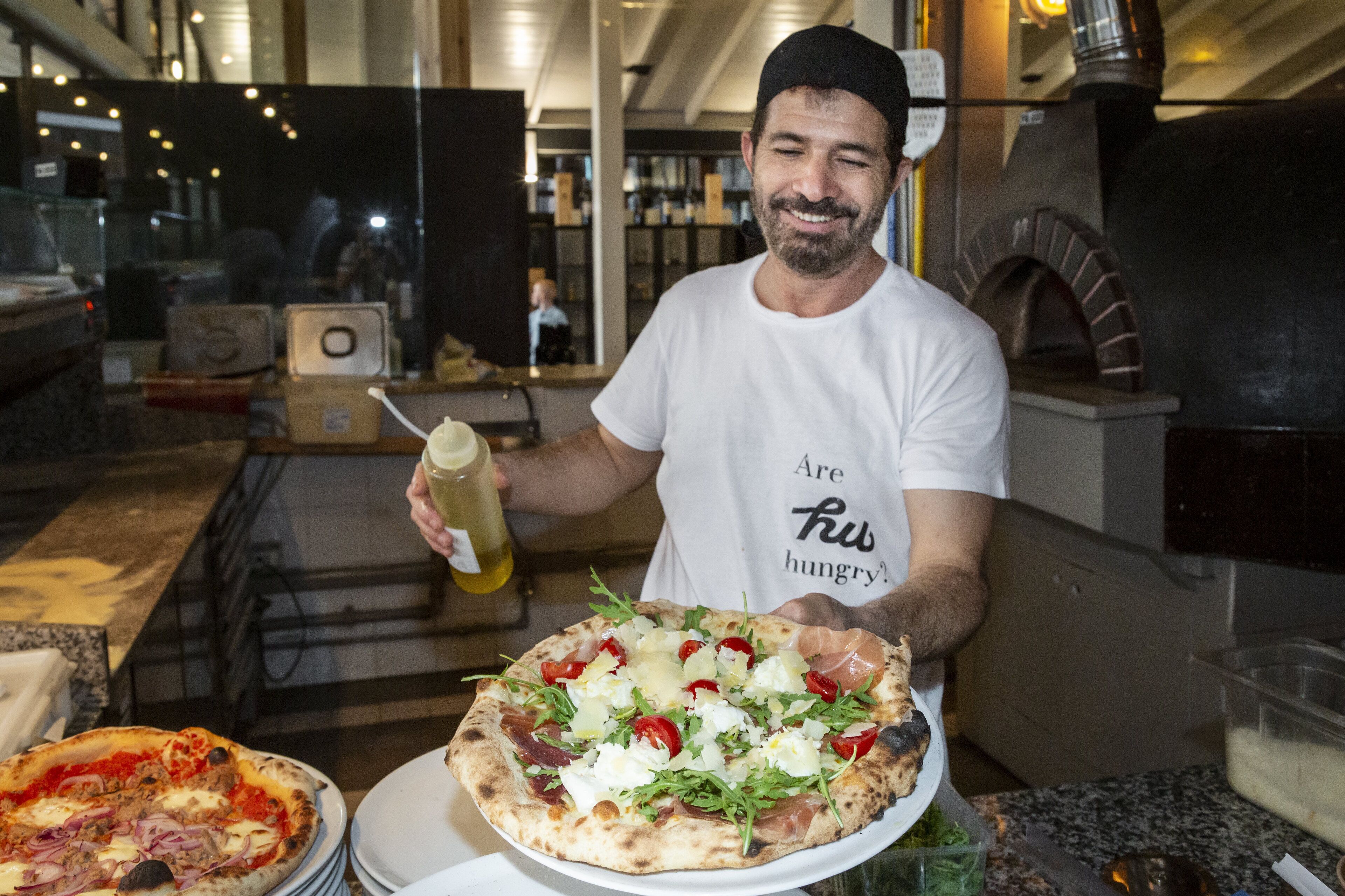 A person holding a pizza with various toppings, standing in a kitchen area with a pizza oven in the background.