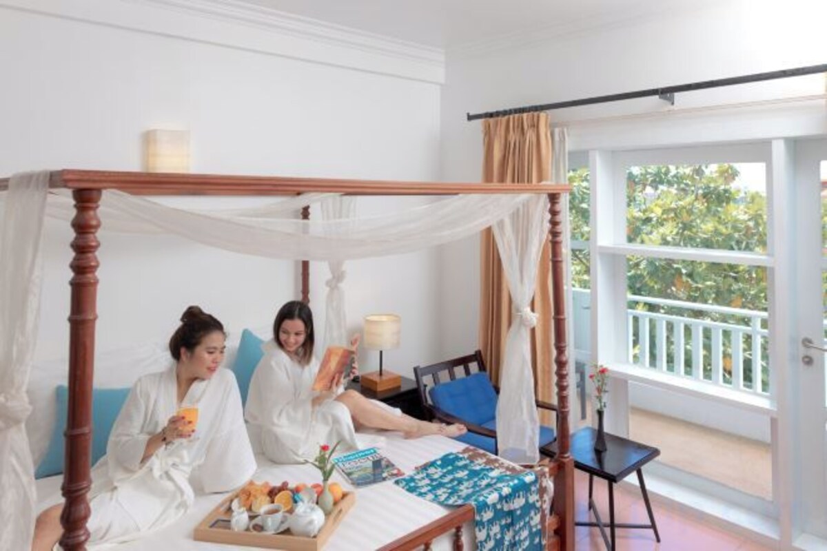 Une chambre avec un lit à baldaquin, une table sur laquelle est posé un plateau de nourriture, et une vue sur l’extérieur grâce à une grande fenêtre.