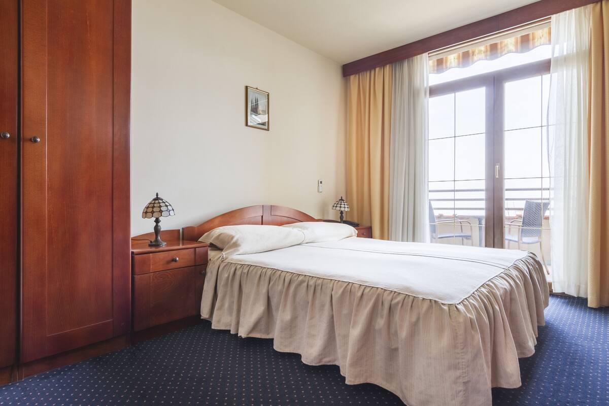 Ein Hotelzimmer mit einem Bett, einem Holzschrank, zwei Nachttischen mit Lampen, einem Fenster mit Vorhängen und einem kleinen Bild an der Wand.
