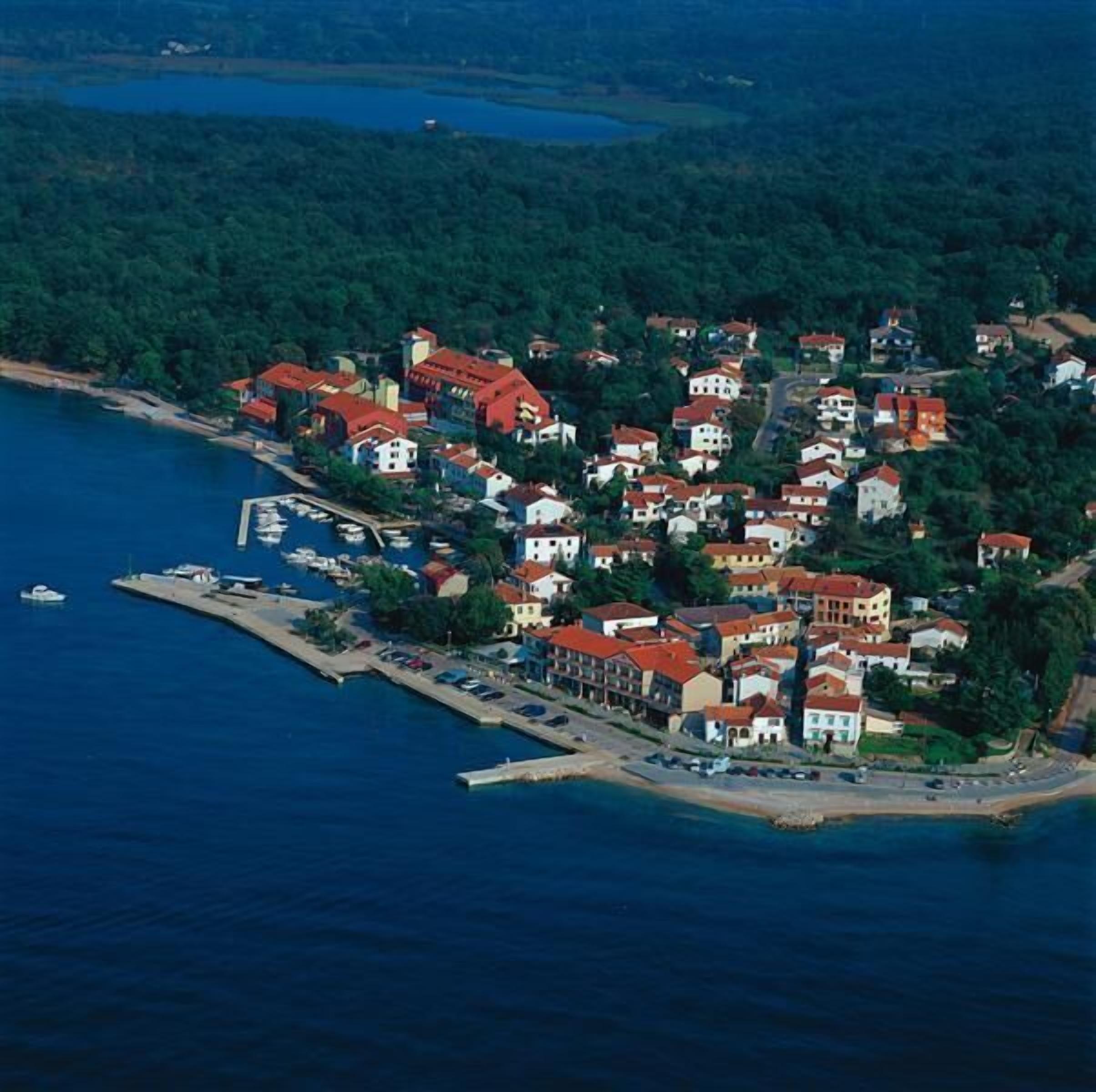 An aerial view of a coastal town with buildings clustered around a marina, surrounded by dense greenery.