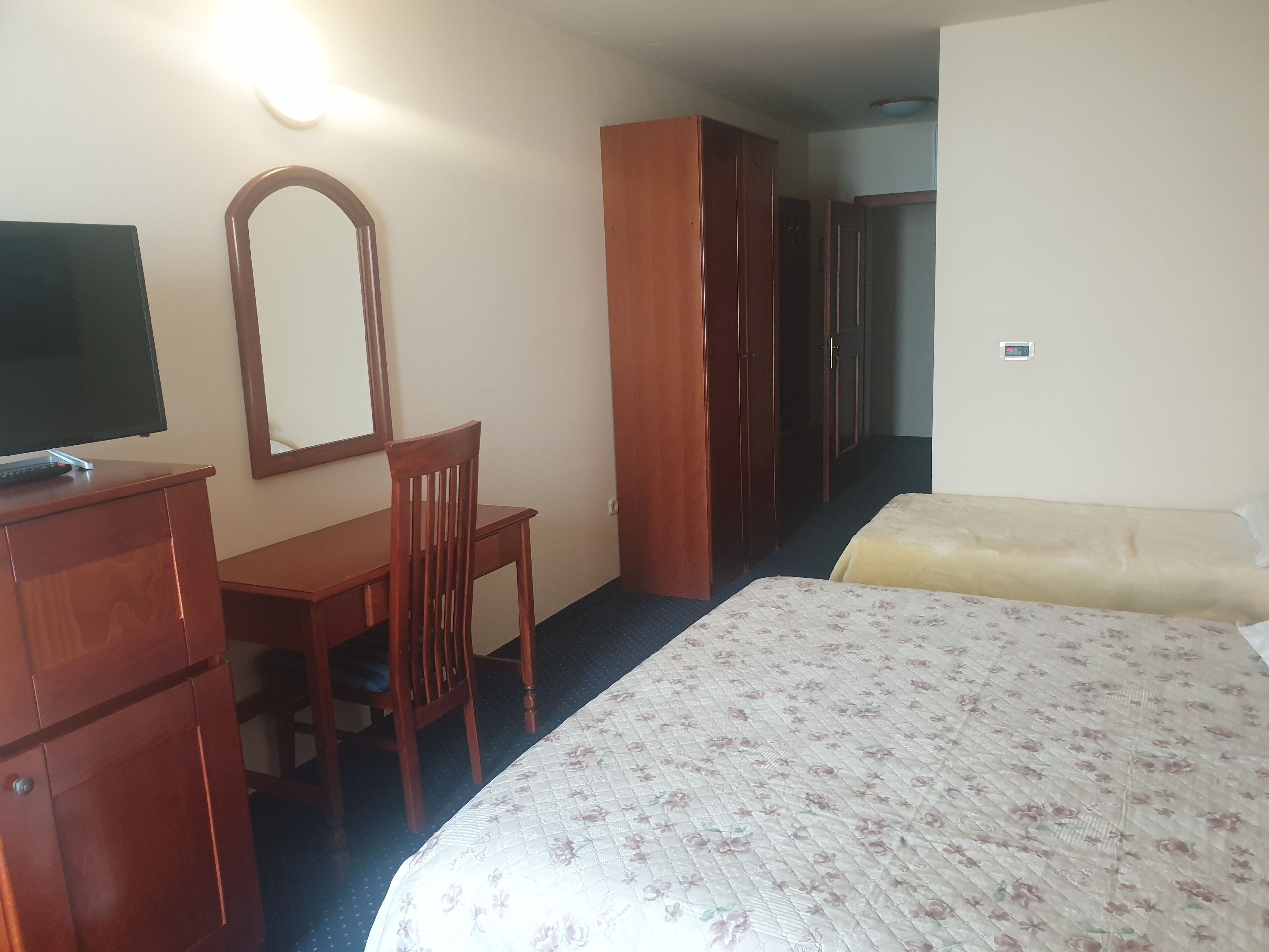 A hotel room with a bed, a desk with a chair, a wardrobe, and a mirror.