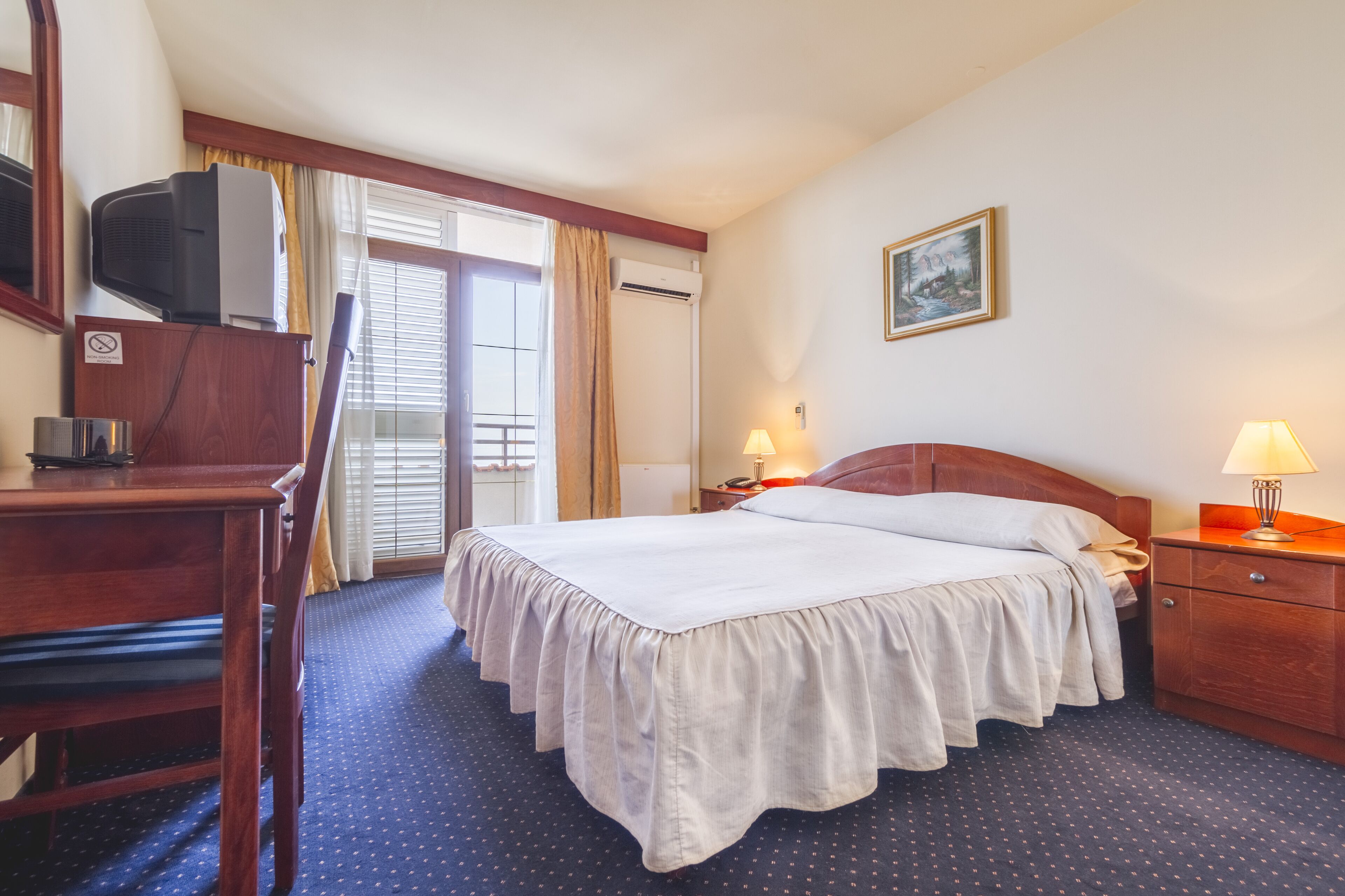 A hotel room with a bed, a desk, a chair, a TV, a lamp, and a painting on the wall.