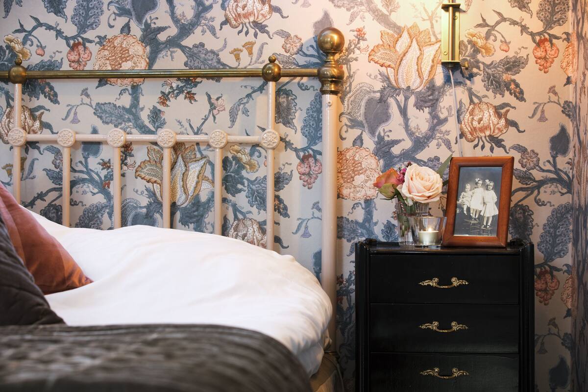 A neatly made bed with a white comforter and a brown pillow, a black nightstand with drawers, a framed picture, and a vase with flowers.