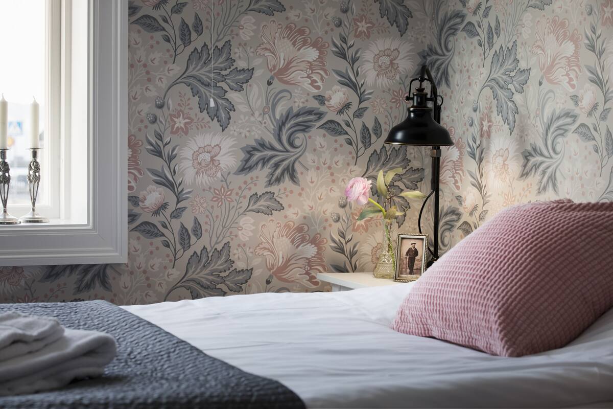 A bedroom with floral wallpaper, a bedside lamp, a pink pillow, and a window with two candles.
