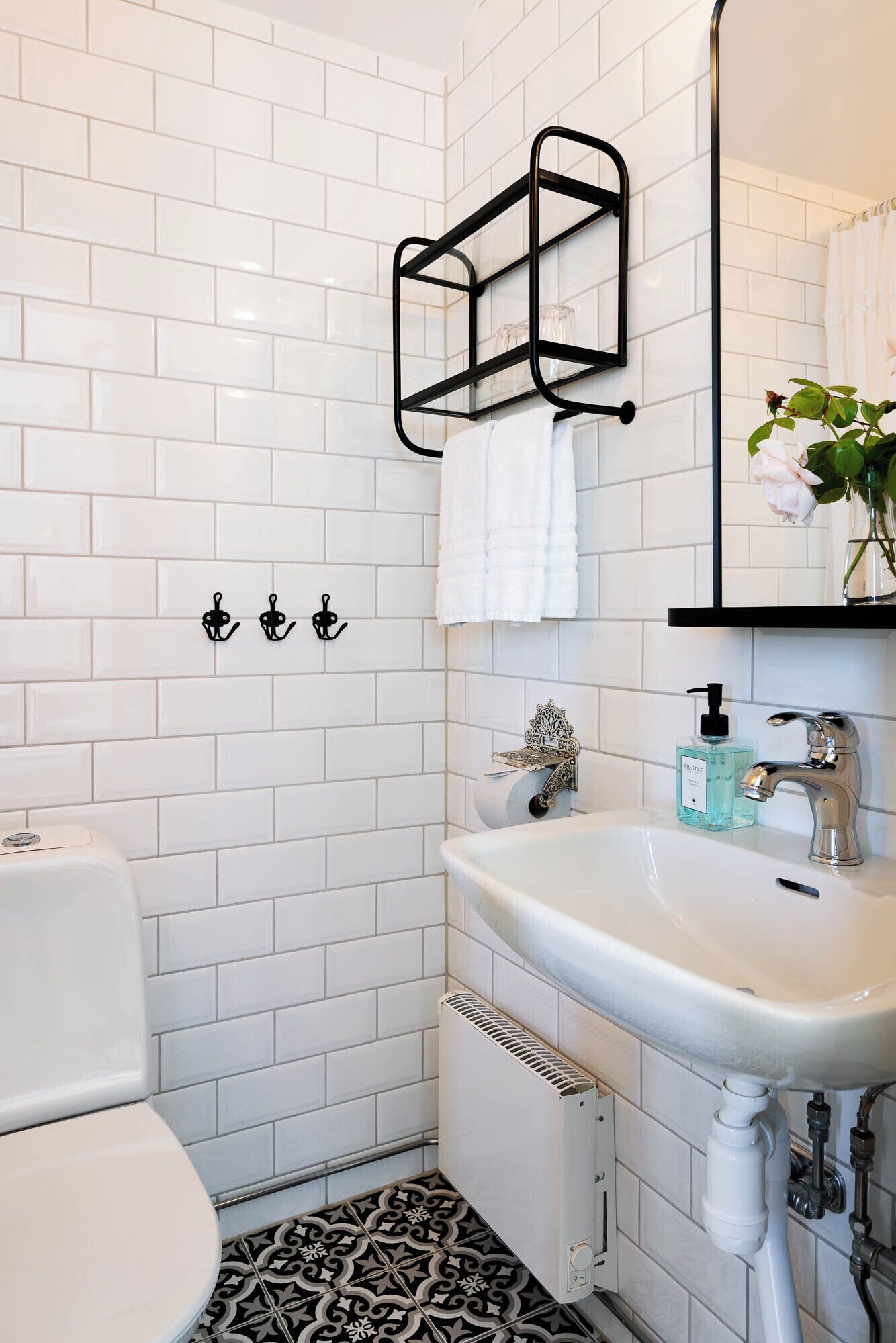 Un baño con azulejos blancos tipo metro, inodoro blanco, piso estampado en blanco y negro, lavabo blanco con grifo plateado, toallero negro y un espejo con un jarrón de flores.