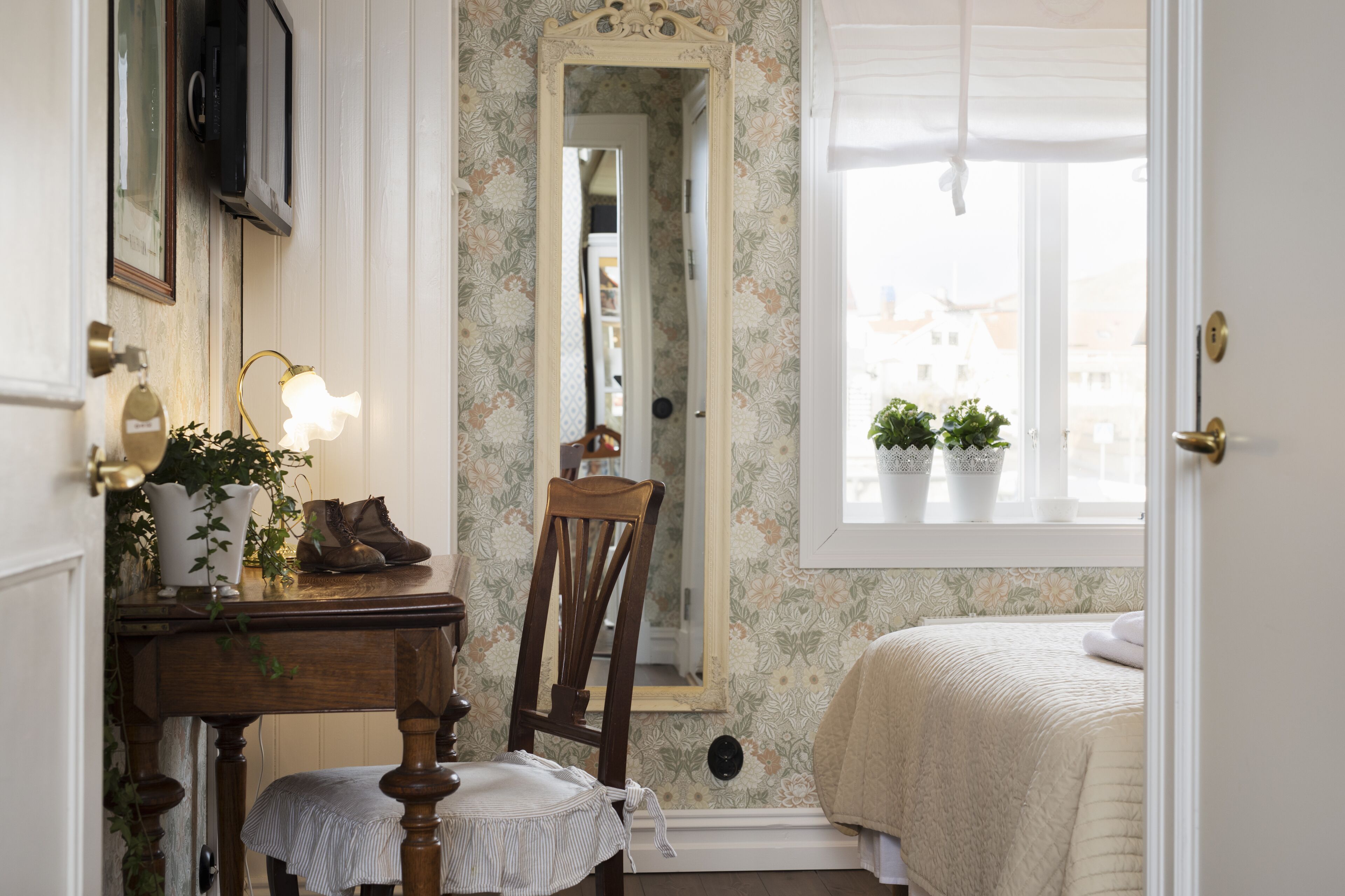 A bedroom with a bed, a chair, a table with a lamp, a mirror, and a window with plants.