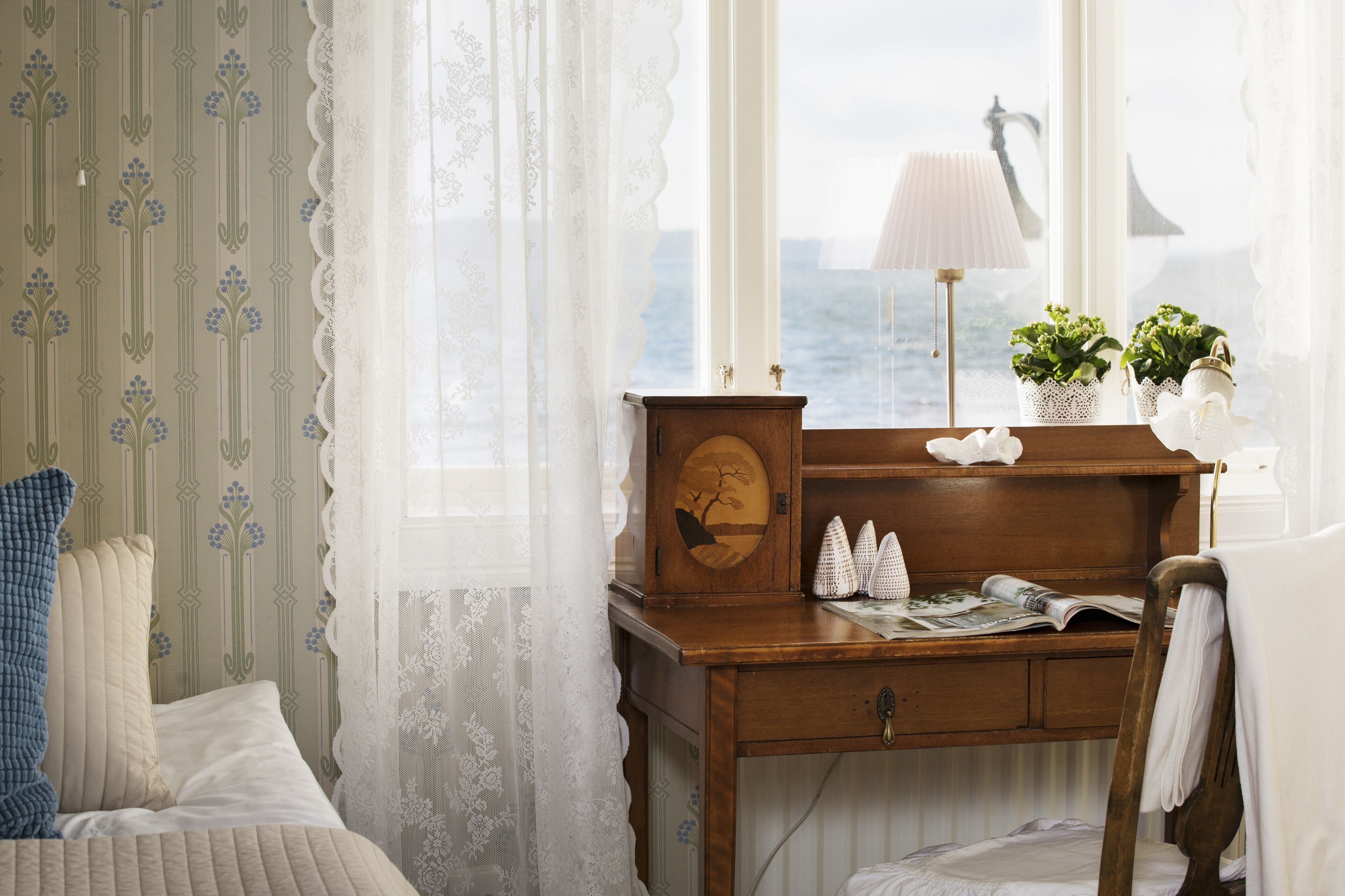 A bedroom with a wooden desk, a bed with white bedding, lace curtains, and a view of the sea through a window.