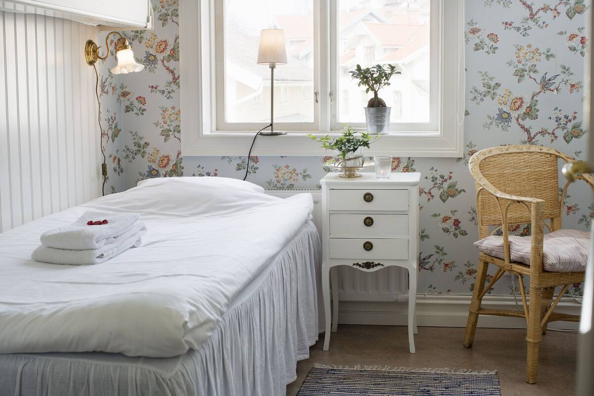 A neatly made bed with white linens and a set of folded towels, a white bedside table with drawers, a wicker chair, and a window with floral wallpaper.