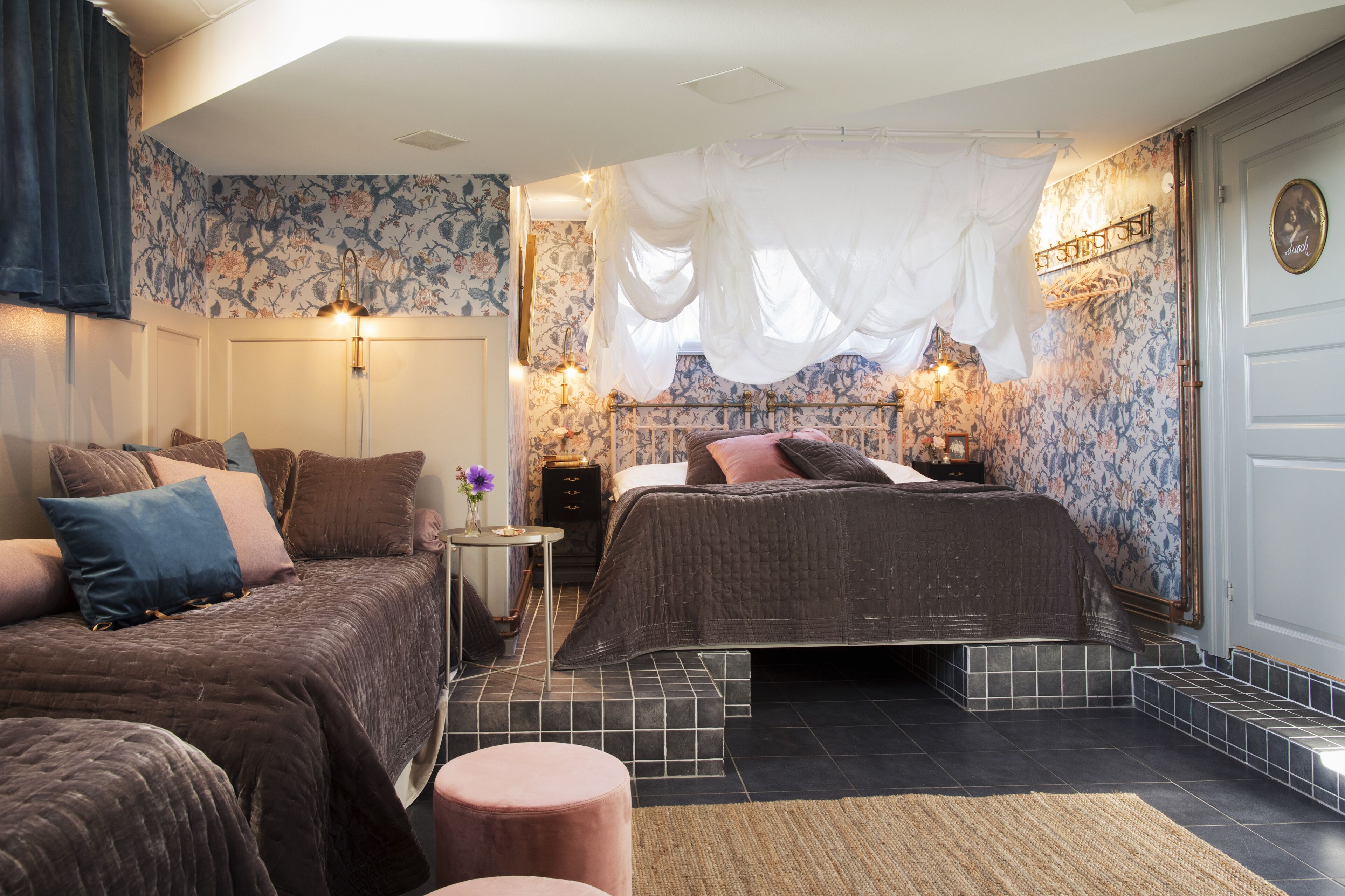Un dormitorio con dos camas, una mesita de noche, una pequeña mesa con un florero y un papel tapiz floral decorativo.