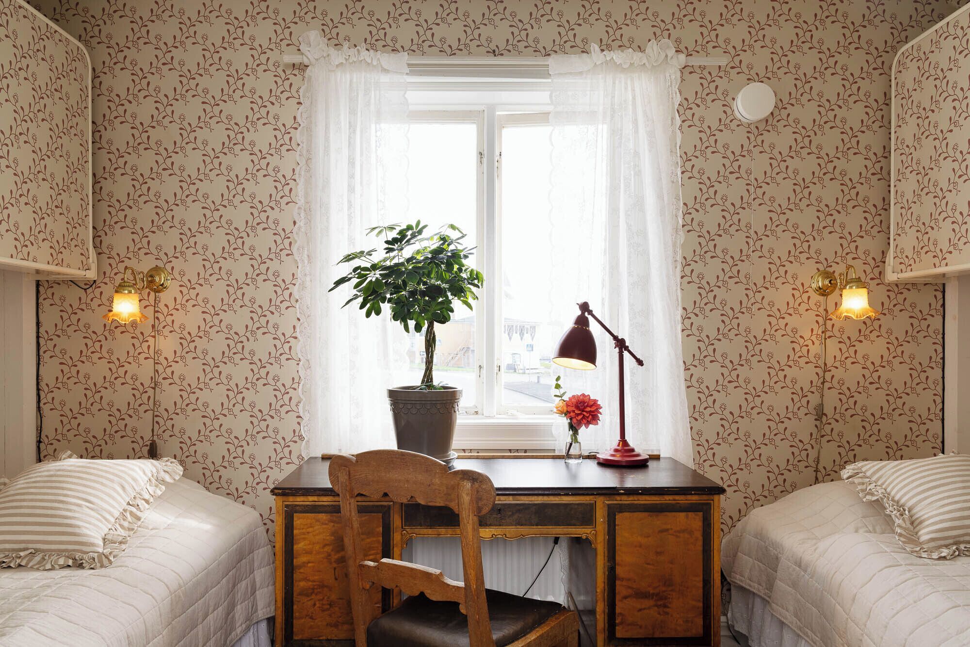 A bedroom with two beds, a desk with a chair, a potted plant, a desk lamp, and a window with a curtain.