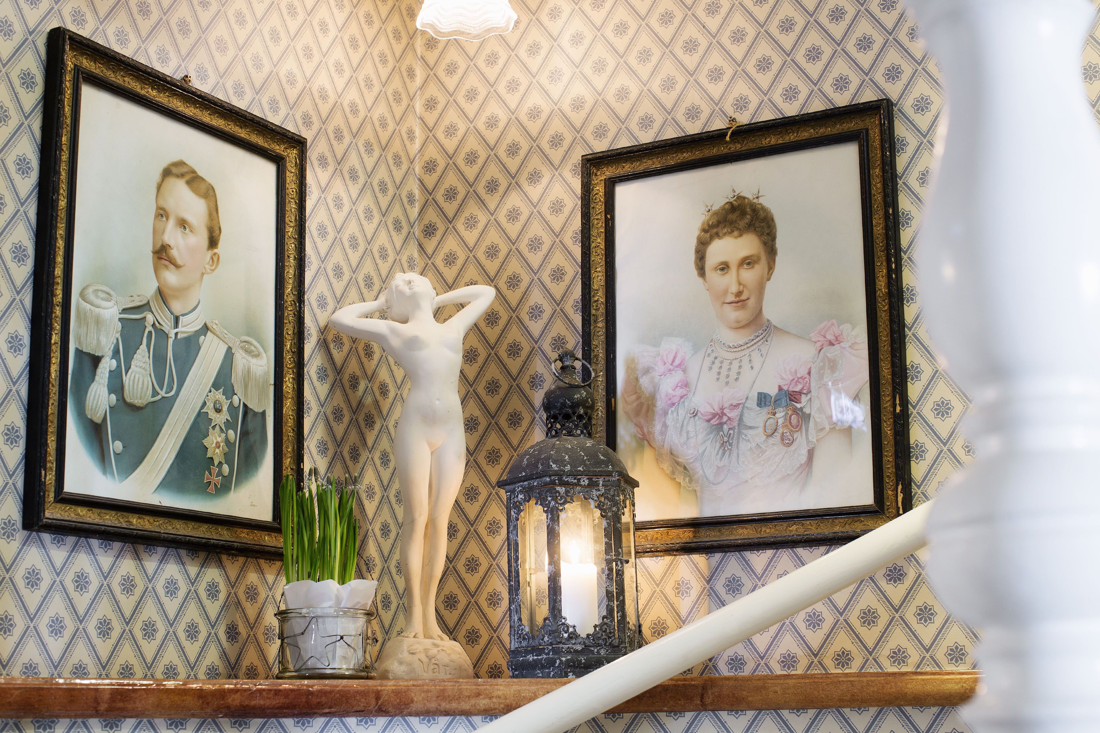 A staircase with a patterned wall, two framed portraits, a white bust, and a lantern.