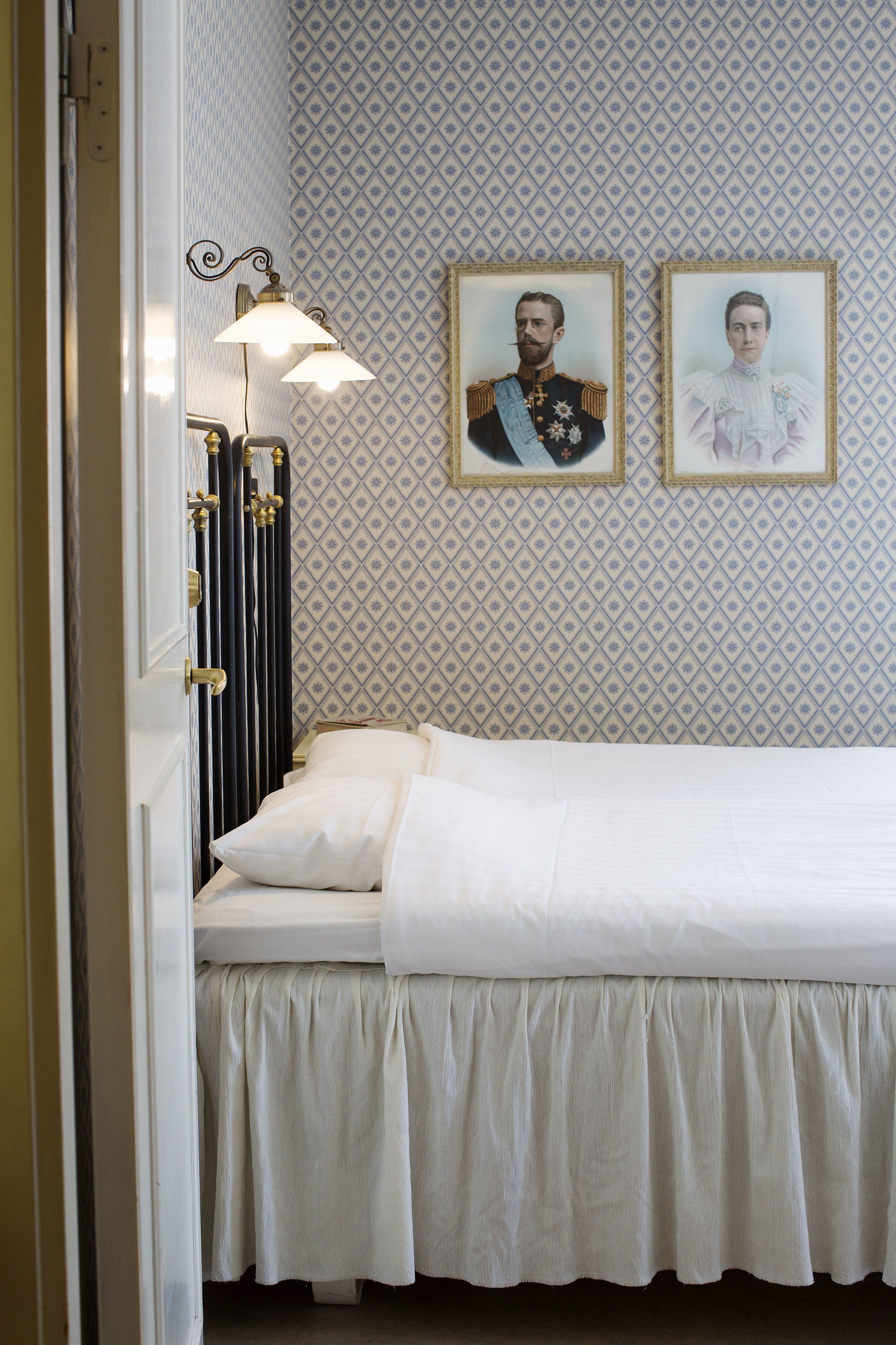 Ein ordentlich bezogenes Bett mit weißer Bettwäsche, eine Wand mit gemustertem Tapeten und zwei gerahmte Portraits darüber.