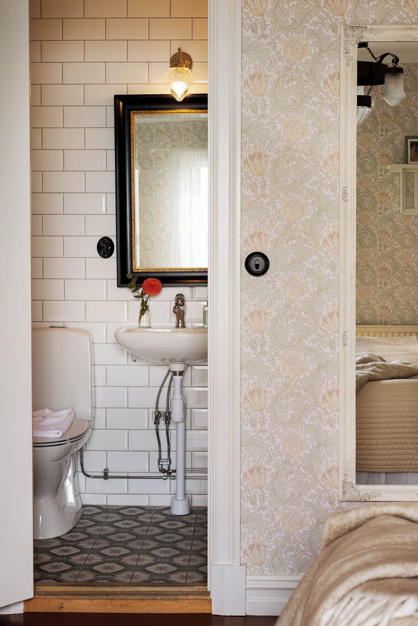 Une salle de bain avec un mur recouvert de carreaux blancs, un lavabo avec un robinet doré, des toilettes et un miroir au-dessus du lavabo.