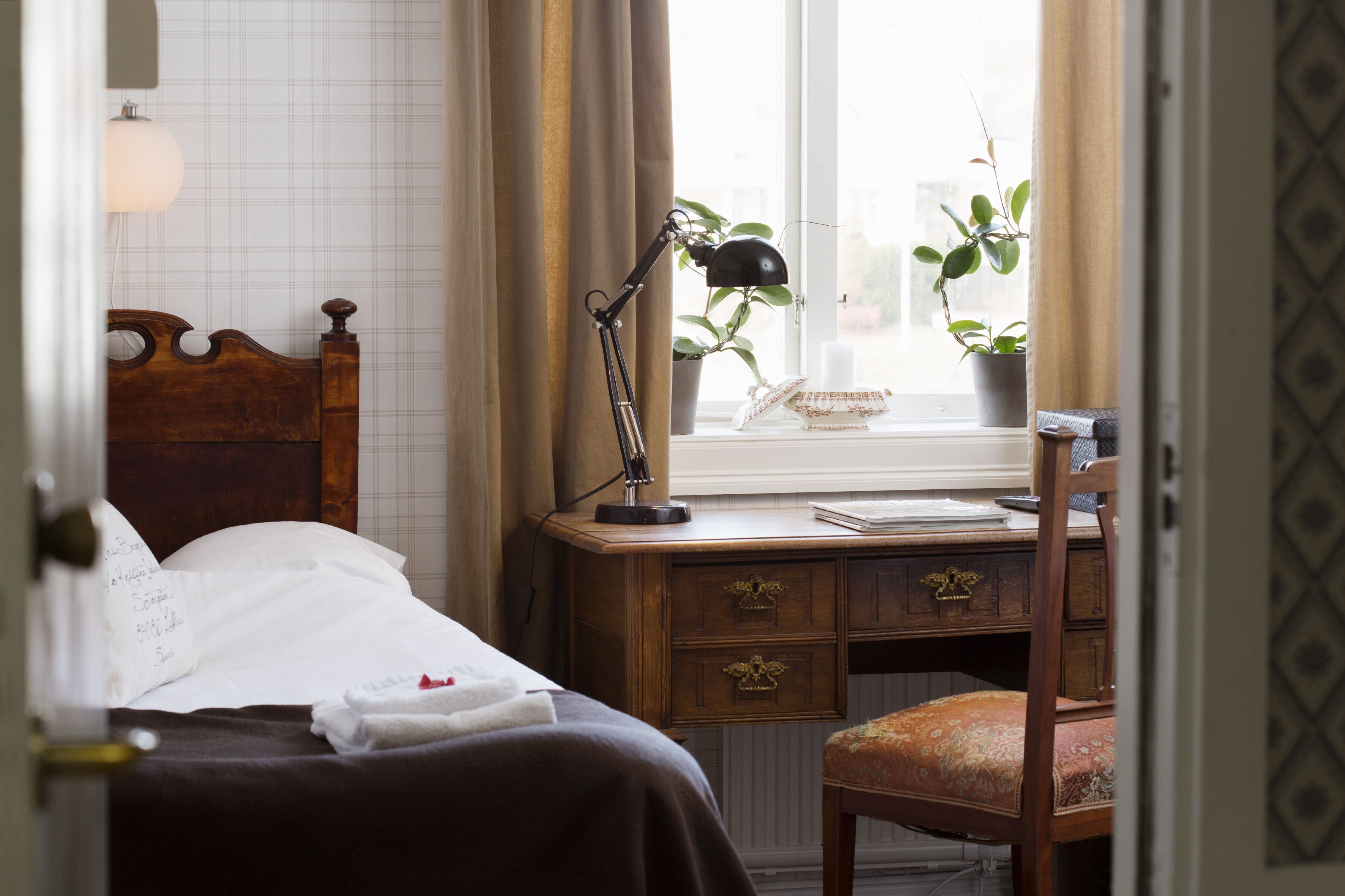 A neatly made bed with a headboard, a desk with a lamp, a chair, and a window with plants.