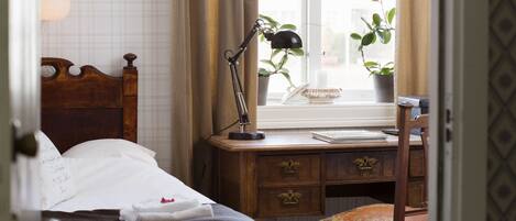 A neatly made bed with a headboard, a desk with a lamp, a chair, and a window with plants.