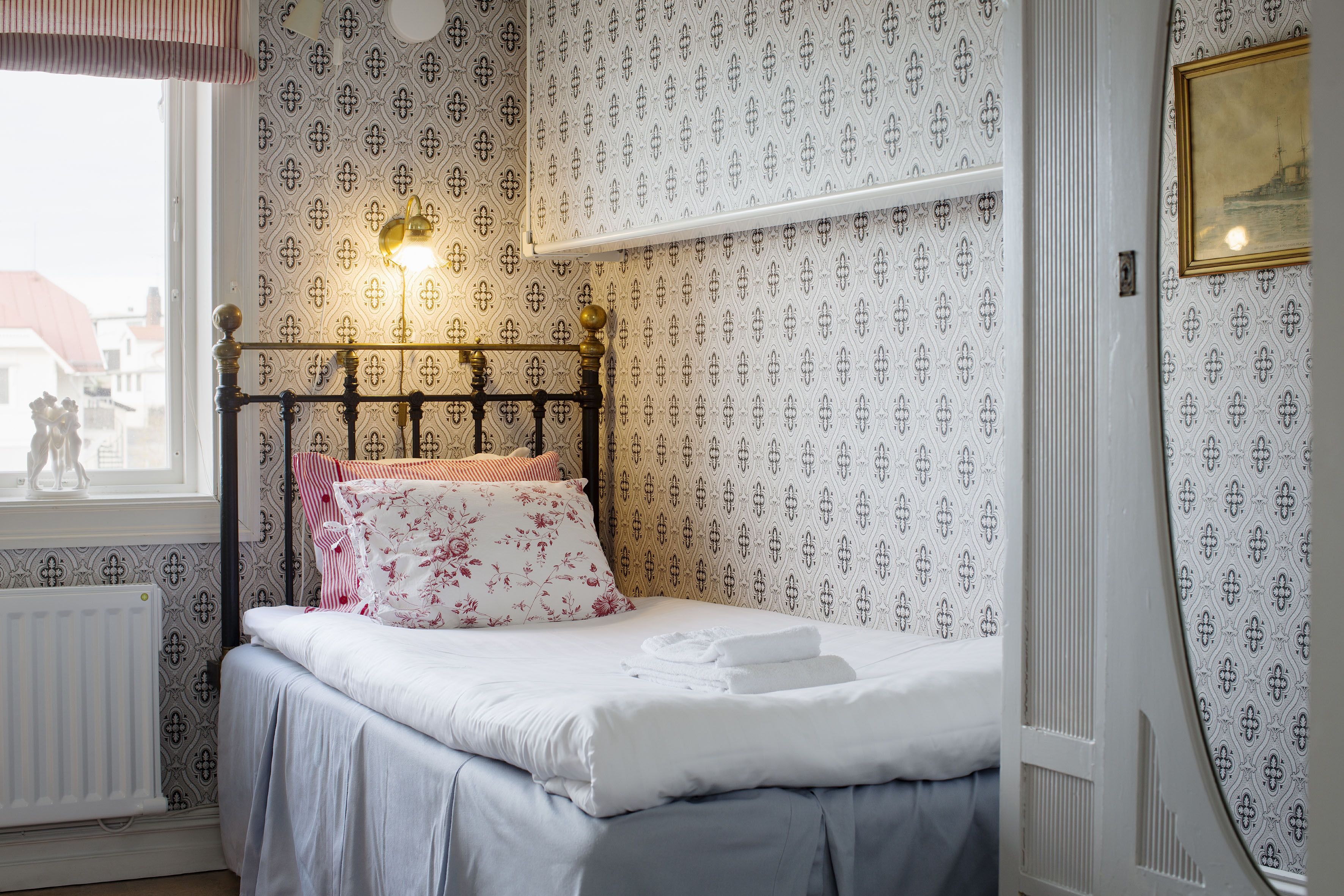 A neatly made bed with white linens and floral pillows, a wall-mounted shelf above, and a window with a view of buildings.