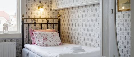 Una cama bien hecha con sábanas blancas y almohadillas estampadas, un estante en la pared y una ventana con vistas a edificios.