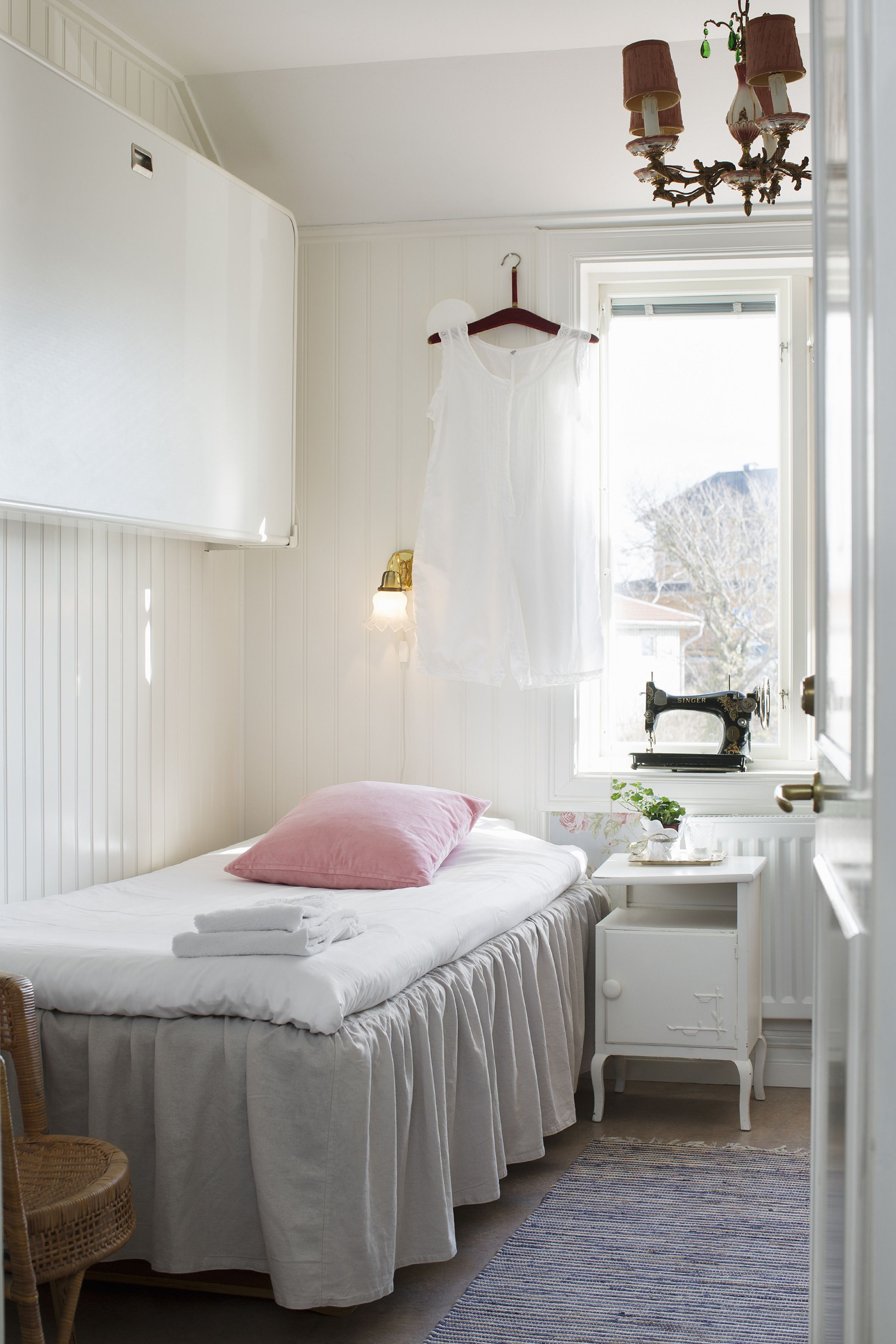 A bedroom with a bed, a dresser, a hanging garment, a sewing machine, and a window with a view.