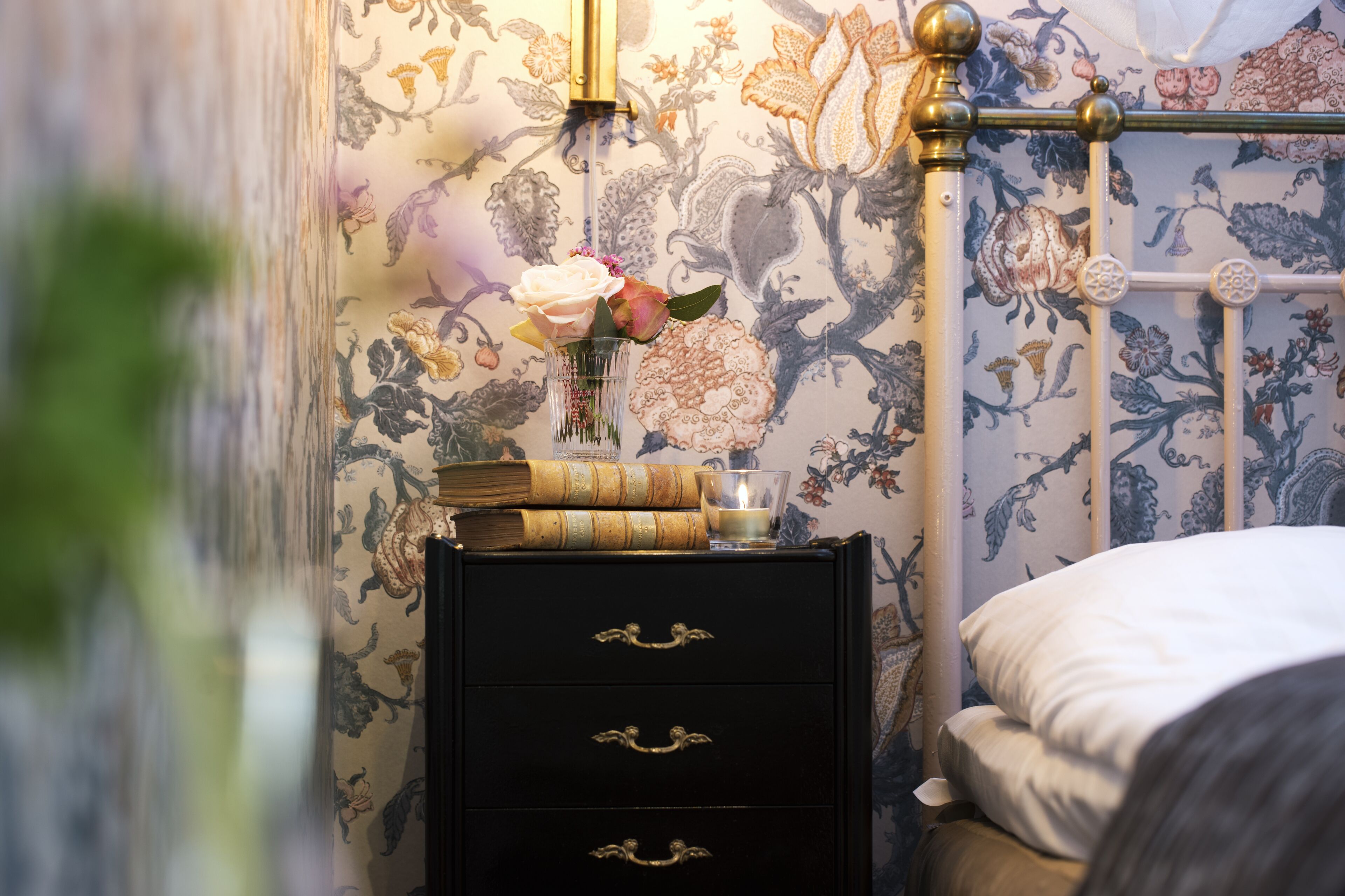Een slaapkamer met bloemenbehang, een nachtkastje met boeken en een kaars, en een bed met wit beddengoed.