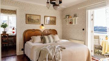 A bedroom with a large bed, a desk with a chair, a potted plant, and a view of the outdoors.