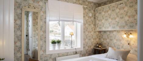 A bedroom with floral wallpaper, a large mirror, a bed with a white comforter, a bedside table with a lamp, and a window with a view of buildings outside.
