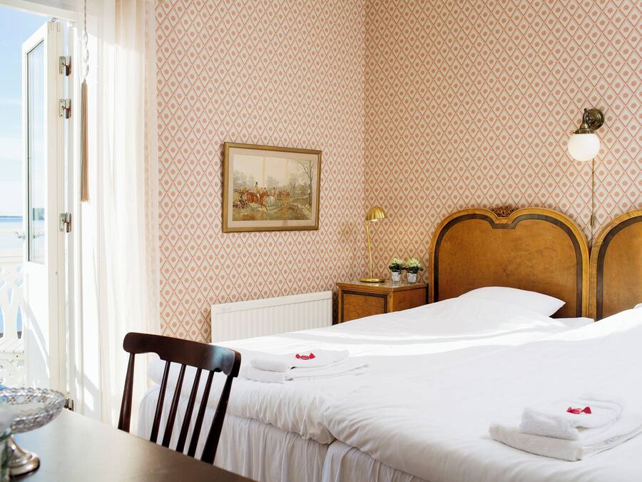 Una habitación de hotel con una cama, una silla, una mesa con un cuenco de vidrio, un cuadro en la pared y vistas al mar a través de una puerta abierta.